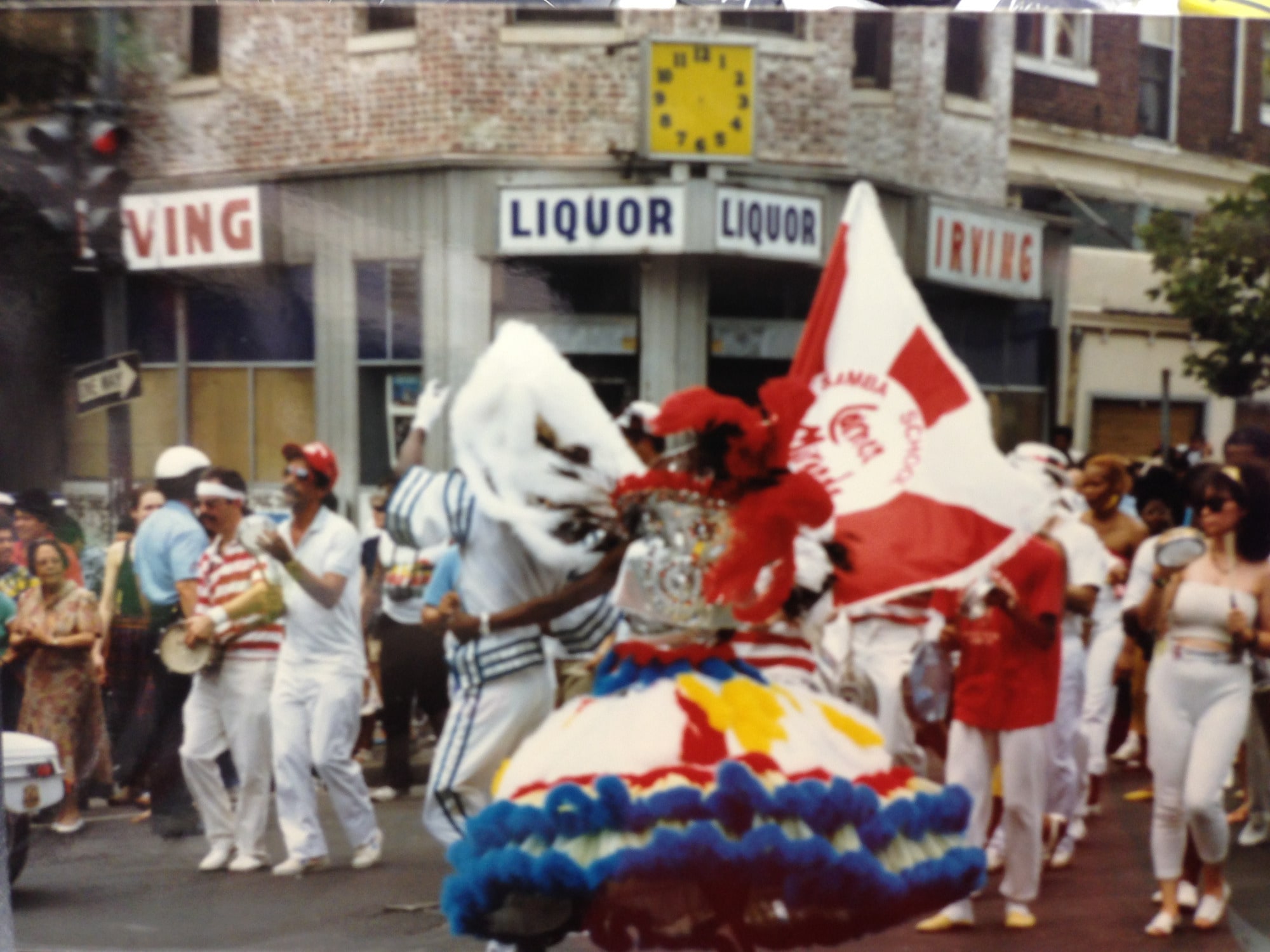 Mount Pleasant Day parade in the 1980s in front of Irving Wine & Spirits — full 'IRVING LIQUOR' signage on both sides of the corner storefront, the yellow clock above, samba dancers in costume in the street.