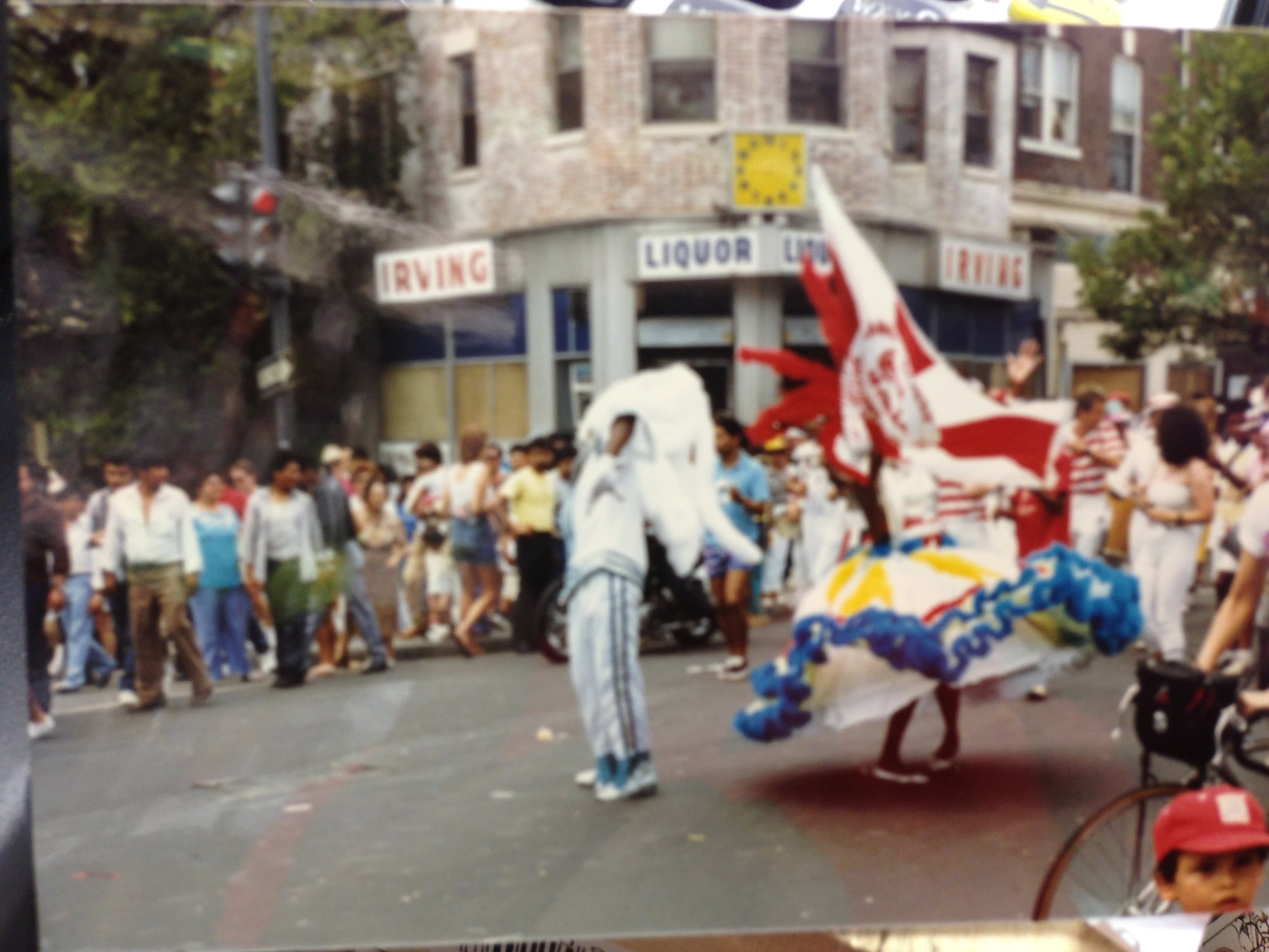 Wide 1980s view of the Mount Pleasant Day parade outside Irving Wine & Spirits — original 'IRVING LIQUOR IRVING' signage, the yellow corner clock, costumed dancers and crowd filling the street.