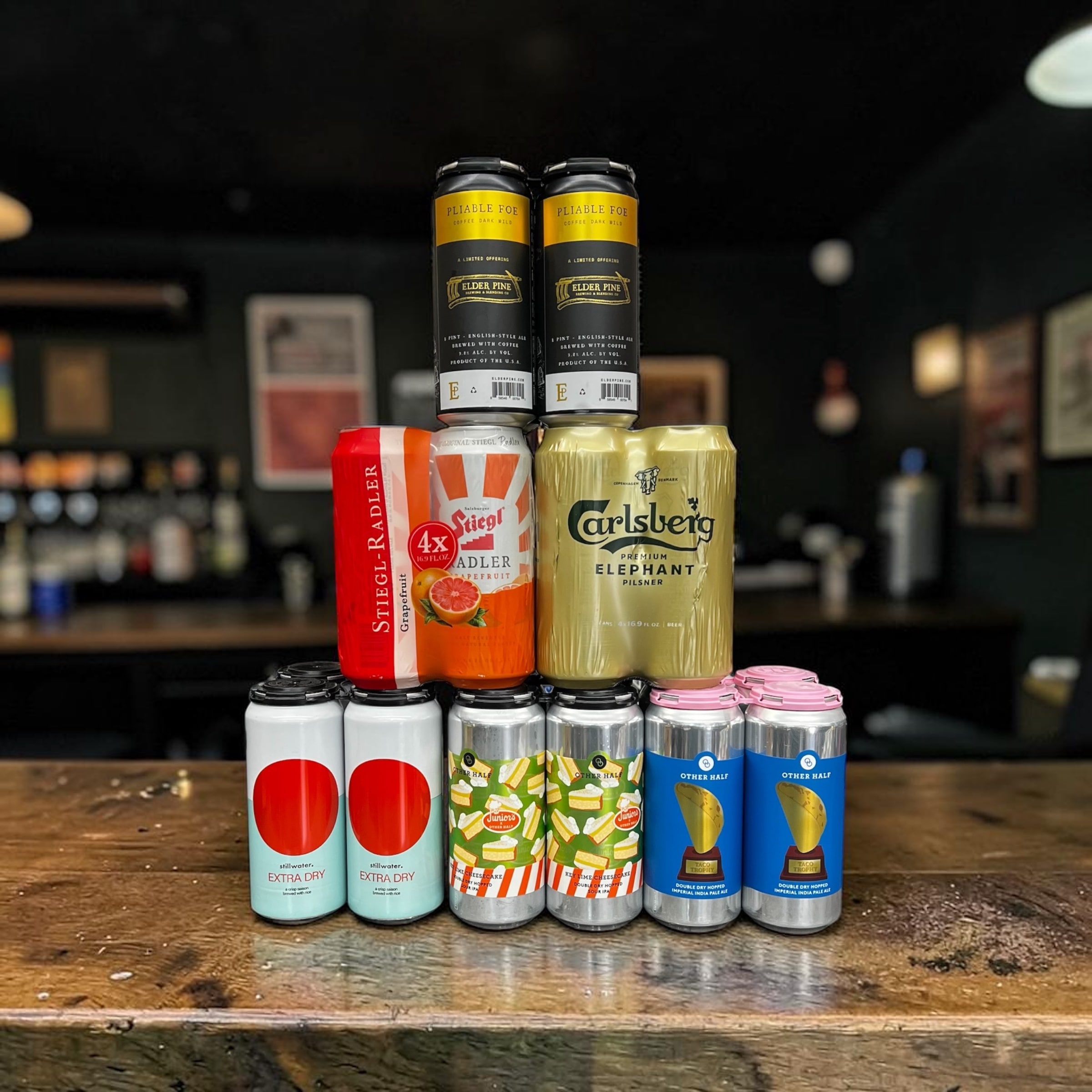 A pyramid of craft beer cans stacked on the counter at Irving Wine & Spirits — Elder Pine on top, Stiegl Radler and Carlsberg Elephant Pilsner in the middle, and Stillwater Extra Dry and Other Half doubles along the bottom.