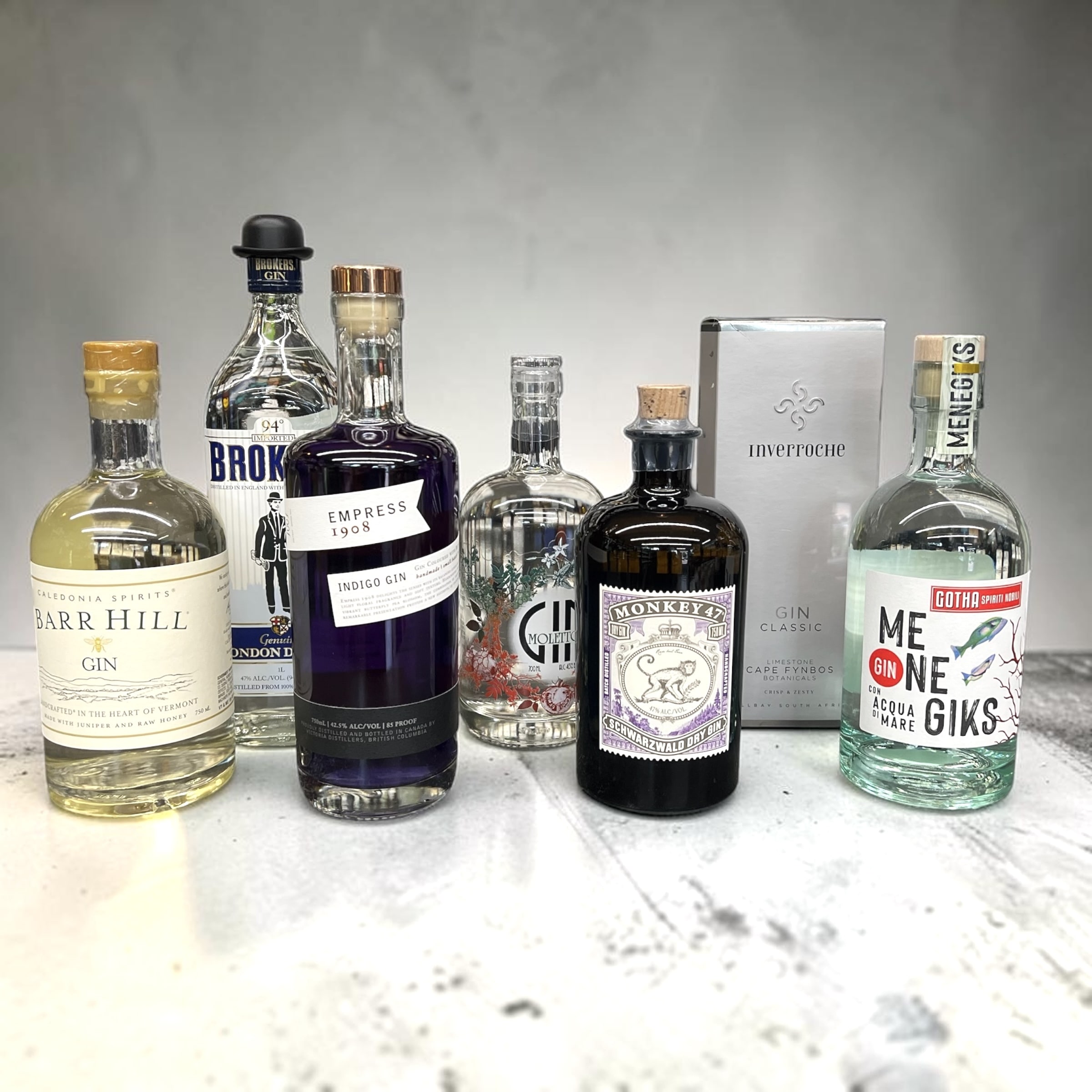 A counter lineup of gins from Irving Wine & Spirits — Barr Hill from Vermont, Brokers London Dry, Empress 1908 indigo gin, Monkey 47 from the Black Forest, Inverroche Cape Fynbos, and Menegik's 'Acqua di Mare' from Italy — every bottle carrying something of where it came from.