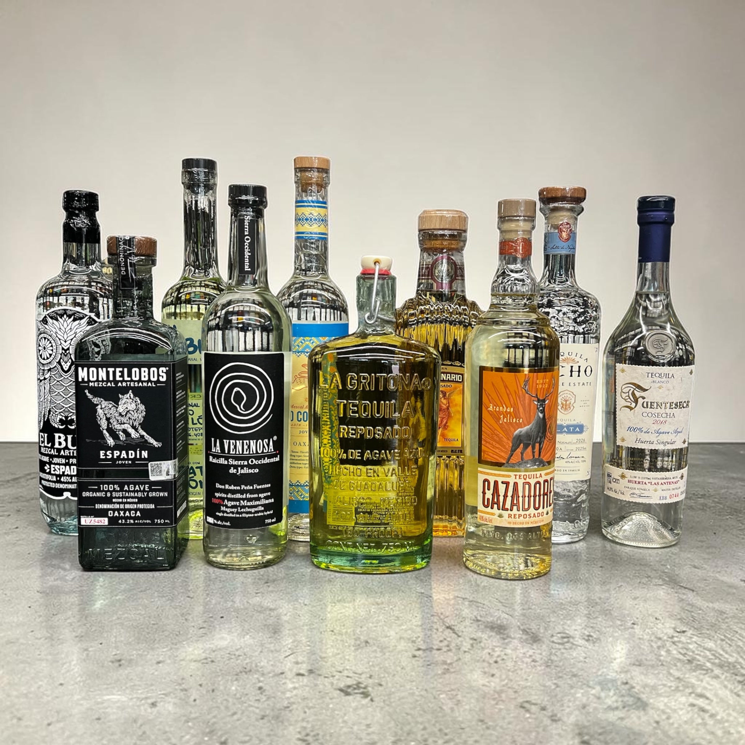 A counter lineup of mezcal and tequila bottles from Irving Wine & Spirits — El Buho and Montelobos Espadín mezcales, La Venenosa Raicilla Sierra Occidental de Jalisco, 400 Conejos joven mezcal, La Gritona Reposado, Cazadores Reposado, and more — the everyday pours next to the special-occasion bottles.