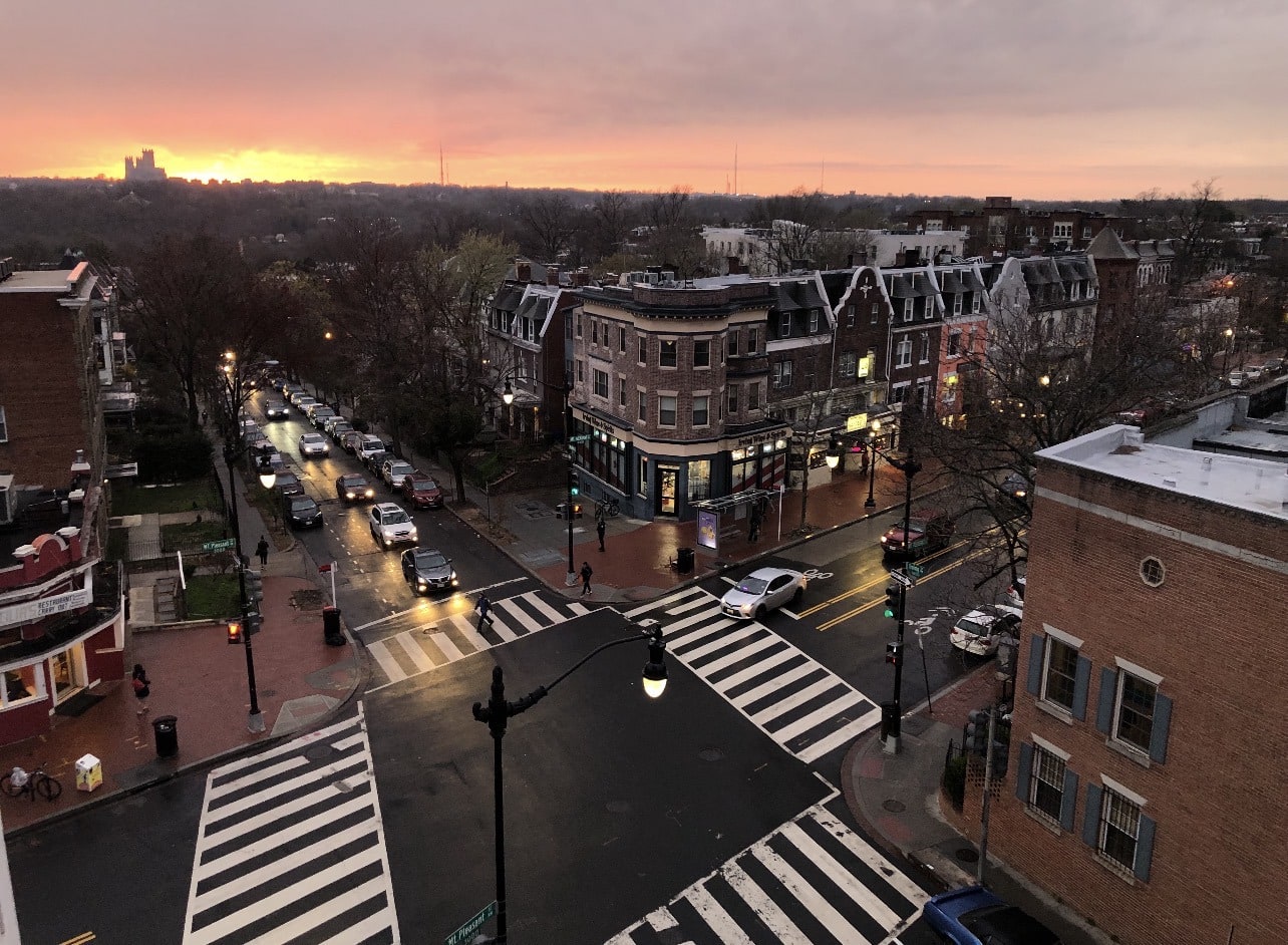 Mount Pleasant Street NW at sunset — wet streets, glowing storefronts, rowhouses catching the last orange light.