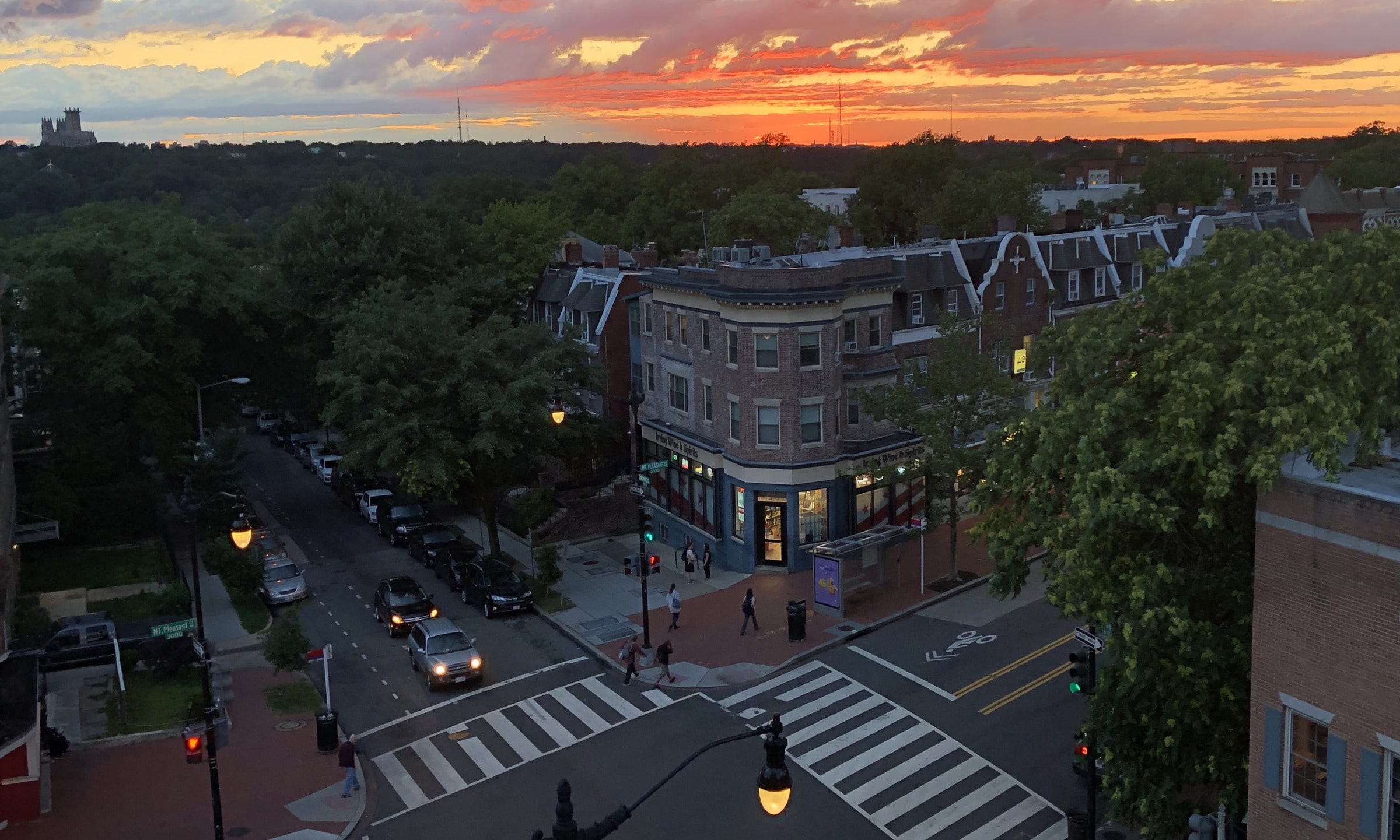 Irving Wine & Spirits at sunset, viewed from across Mt. Pleasant Street NW. The storefront's lights glow on the corner with rowhouses receding into a sunset-orange sky.